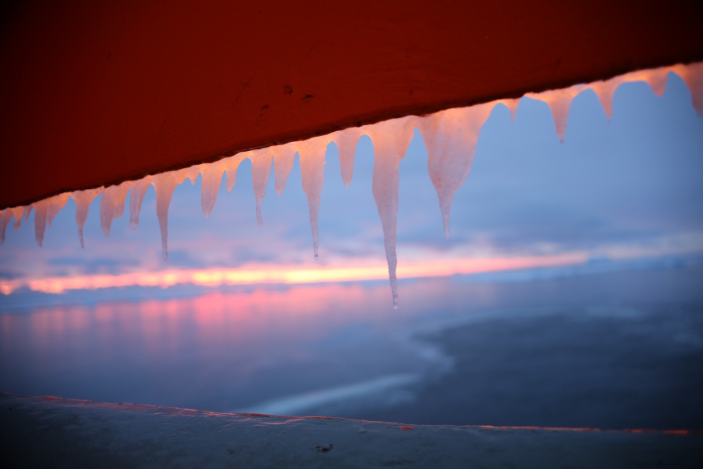 A distant sunset illuminates icicles hanging off a building.